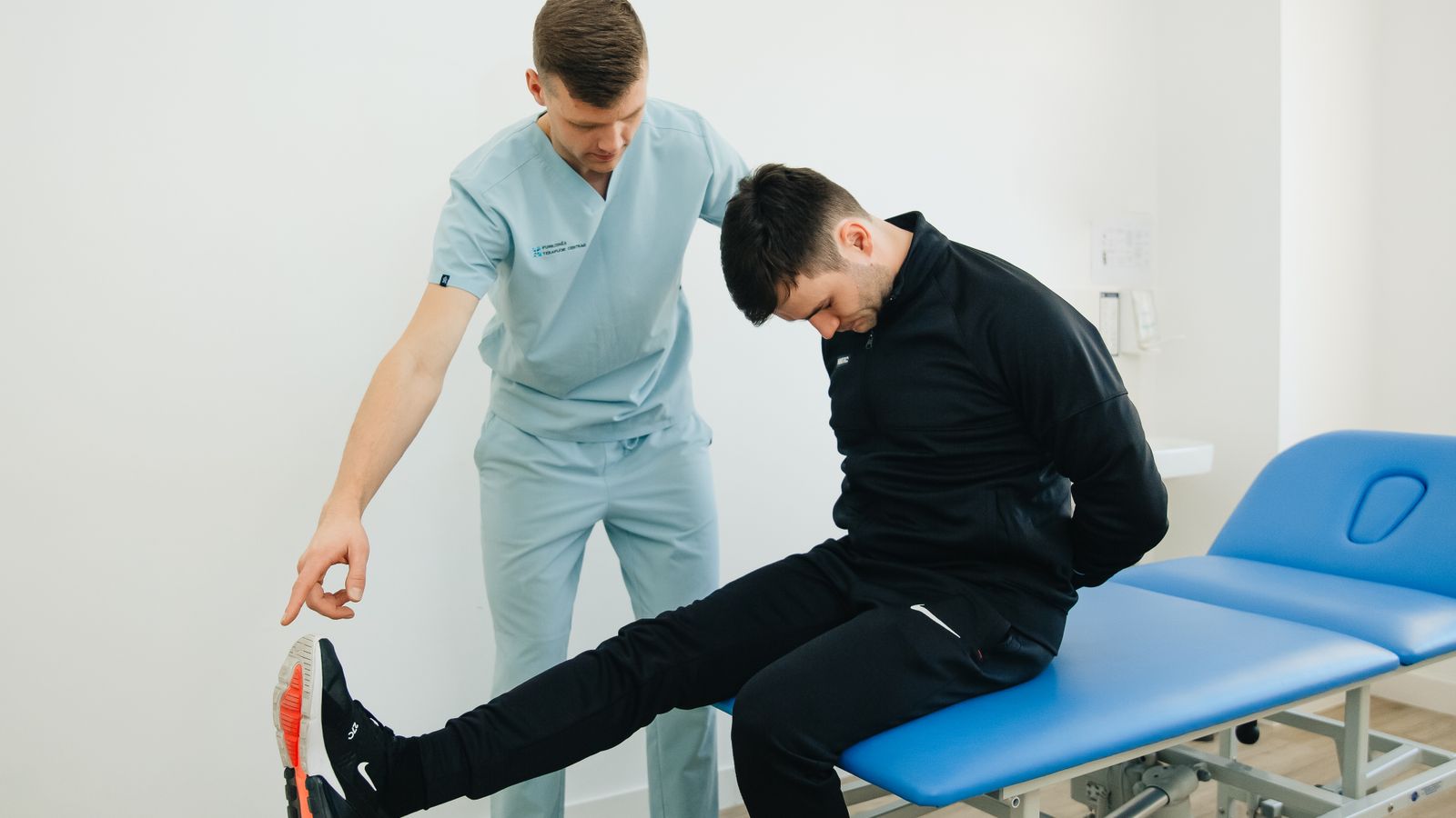 A physical therapist in light blue scrubs guides a seated patient through a hamstring stretch on a blue treatment table