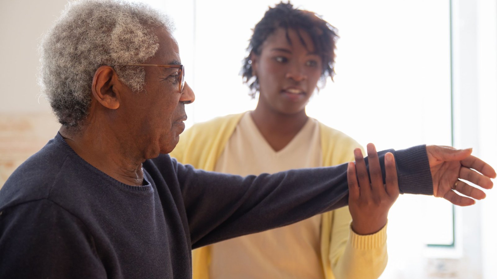 A clinician guides an older man through an arm-raise exercise in a bright room